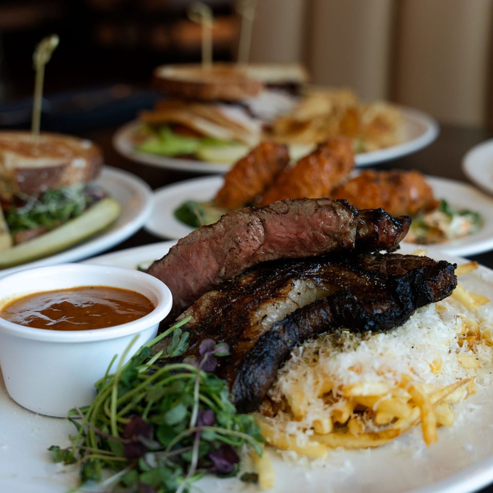 Rivage Oak Kitchen's grilled steak with fries, greens, and dipping sauce, with other dishes blurred in the background.