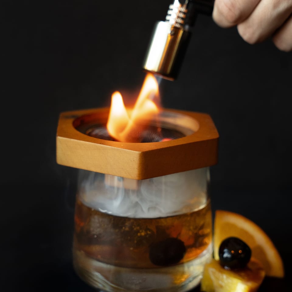 A hand uses a torch to smoke a Rivage Oak Kitchen cocktail, garnished with an orange slice and cherry.