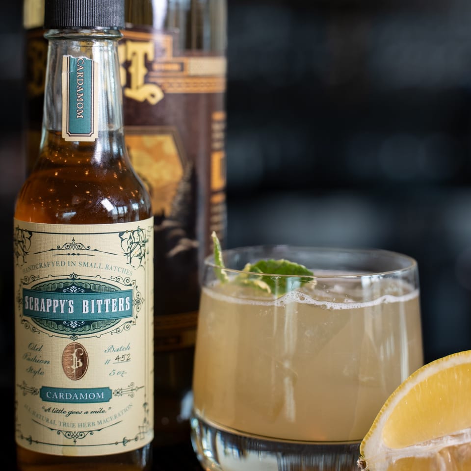 A bottle of Scrappy's Cardamom Bitters next to a cocktail garnished with lime and a lemon wedge.