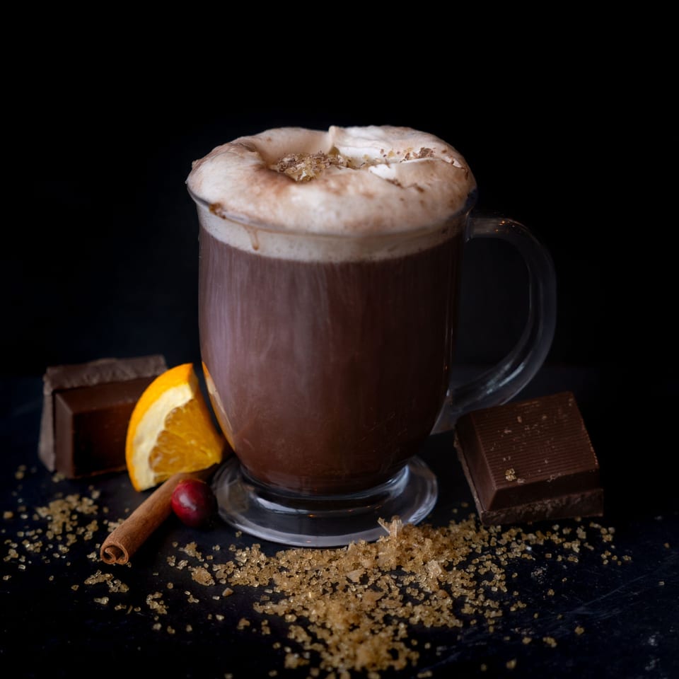 Hot chocolate with whipped cream, orange slice, cinnamon stick, and chocolate squares on a dark surface.