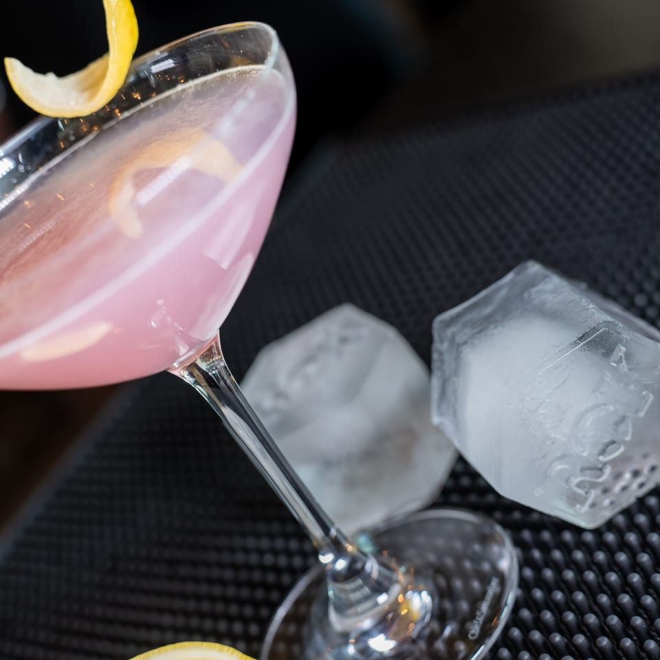 Pink cocktail in a martini glass with a lemon peel garnish, next to large ice cubes.