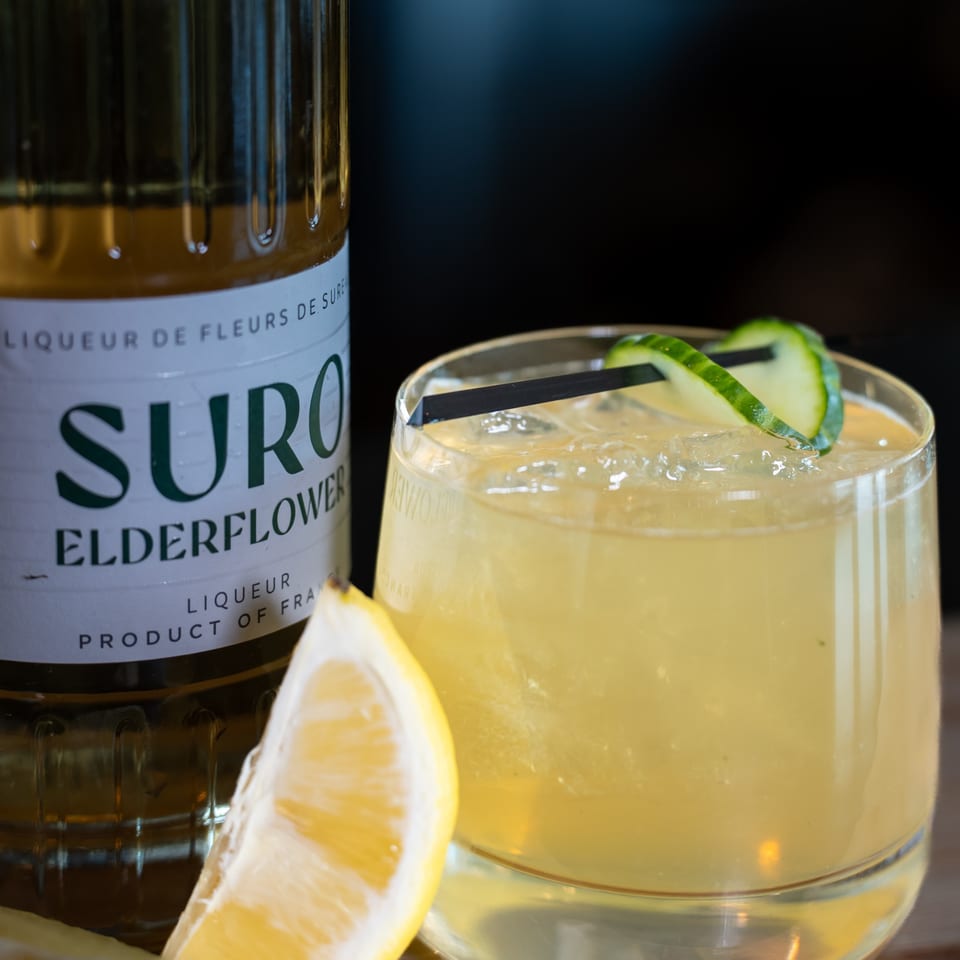 Elderflower liqueur bottle next to a cocktail with a lemon wedge and cucumber garnish.