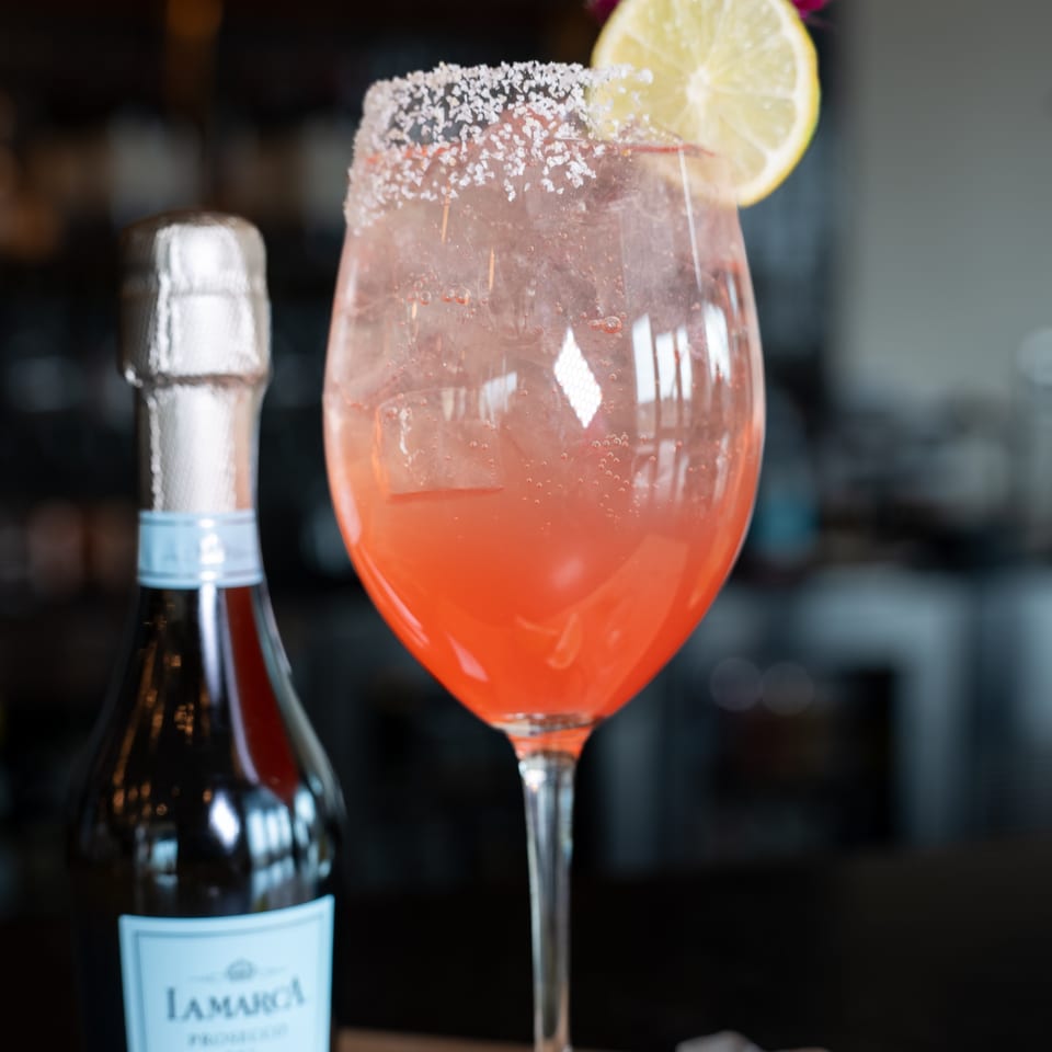 A pink cocktail with a sugared rim and lemon slice next to a mini bottle of La Marca Prosecco.