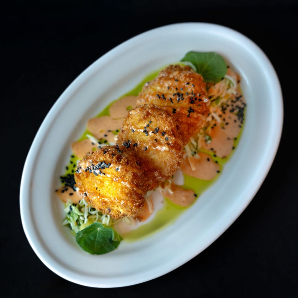 Three golden-fried croquettes with black sesame seeds, drizzled with sauces and garnished with greens.