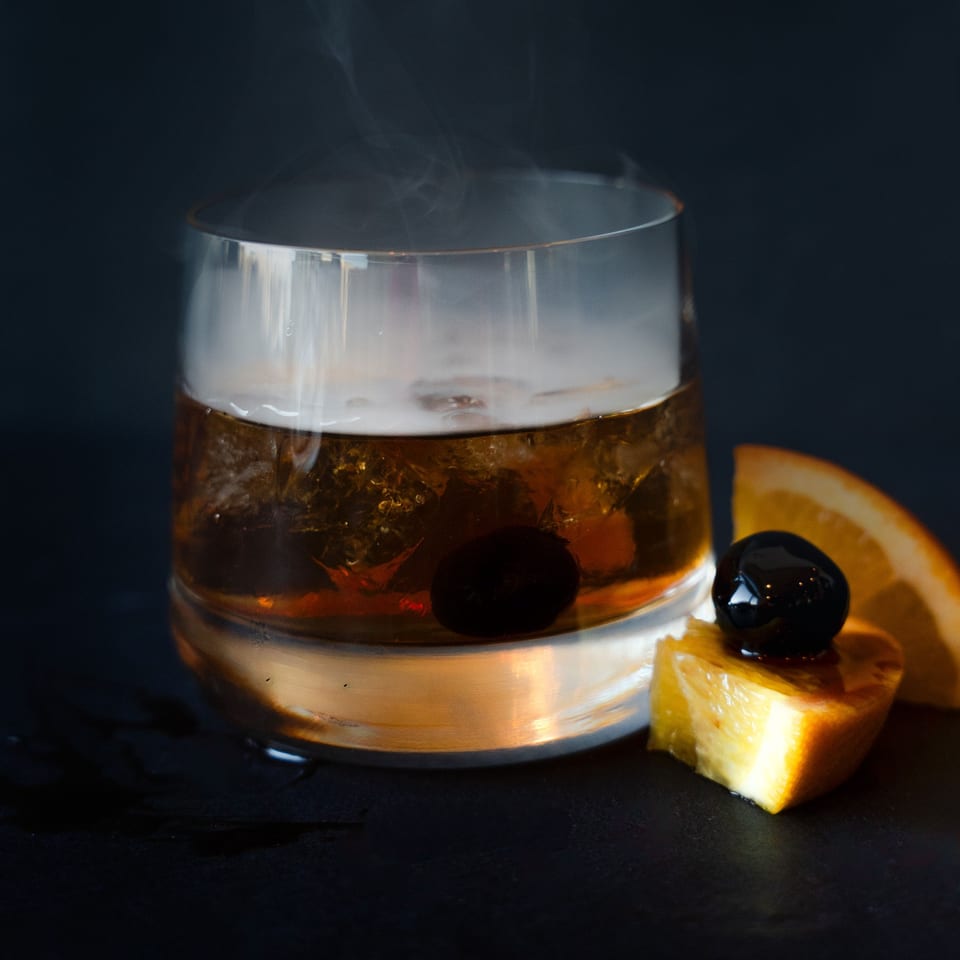 A smoked Old Fashioned cocktail with a cherry and orange slice on a dark surface.
