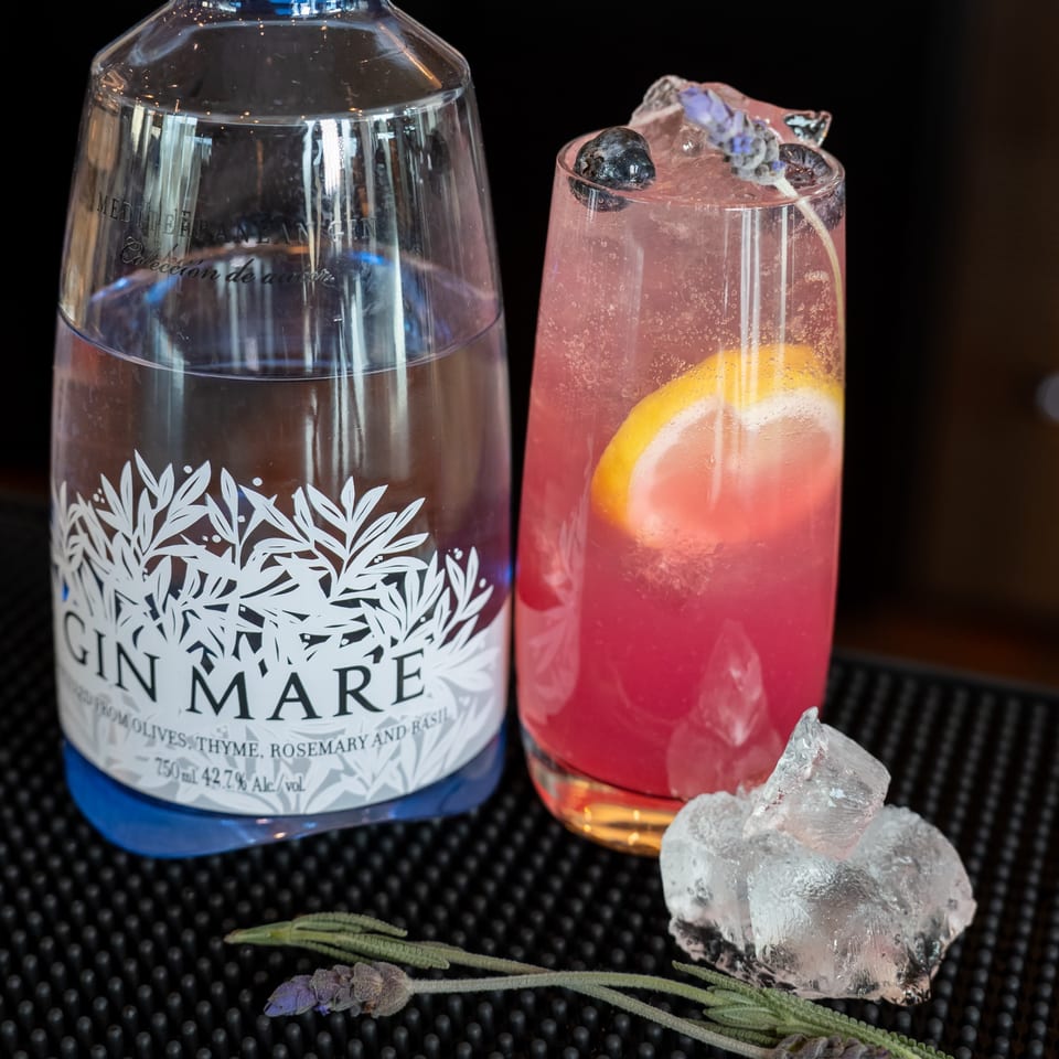 Gin Mare bottle next to a pink cocktail with lemon, blueberries, and lavender on a bar.