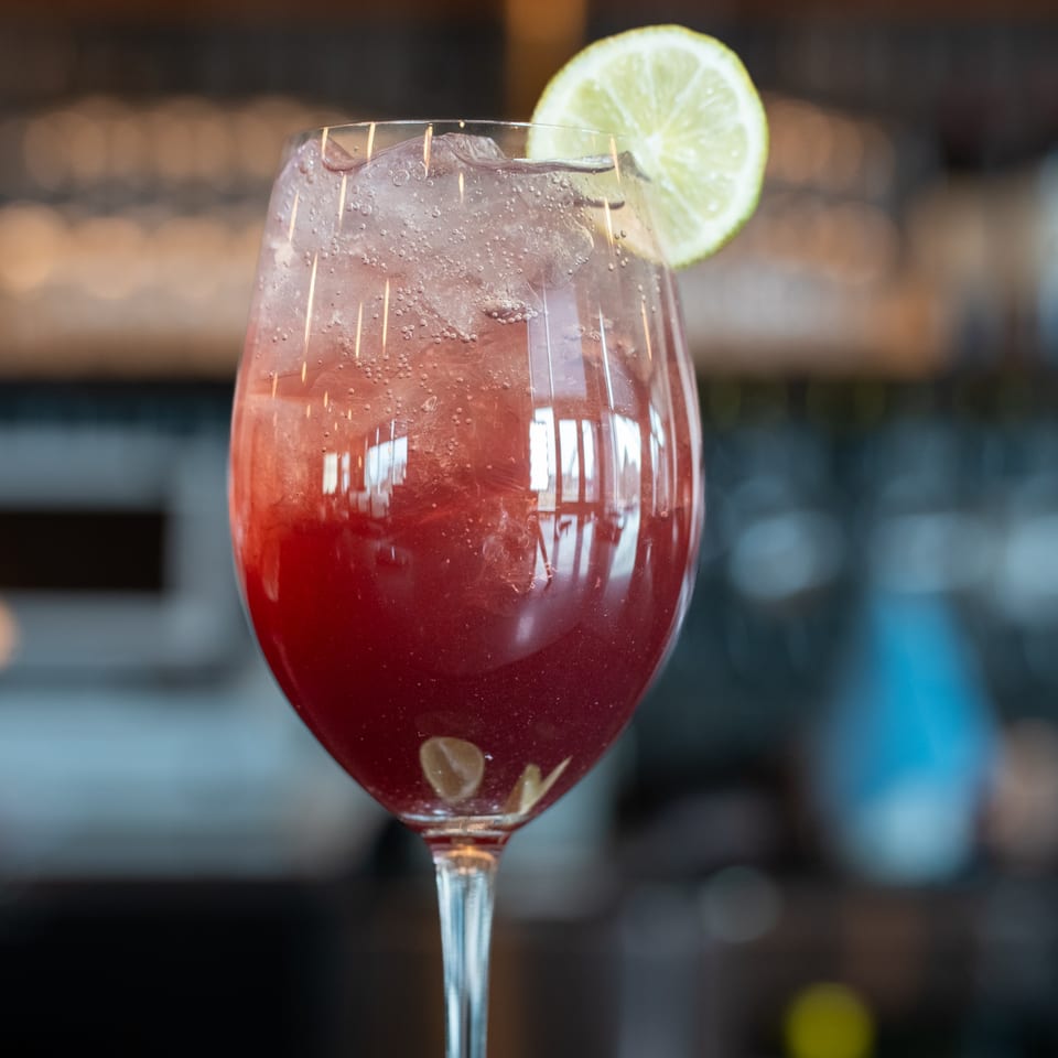 A vibrant red cocktail with ice and a lime slice in a stemmed glass.