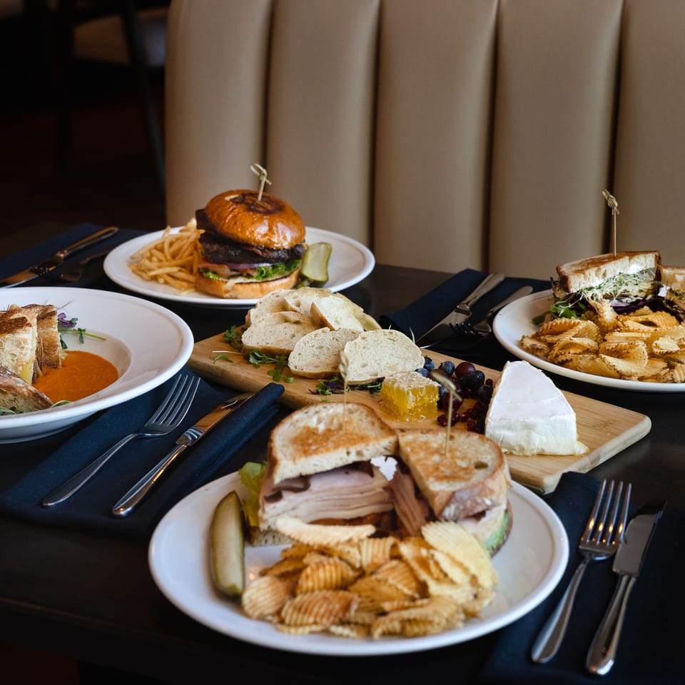 A table at Rivage Oak Kitchen laden with a burger, sandwiches, soup, and a cheese board.